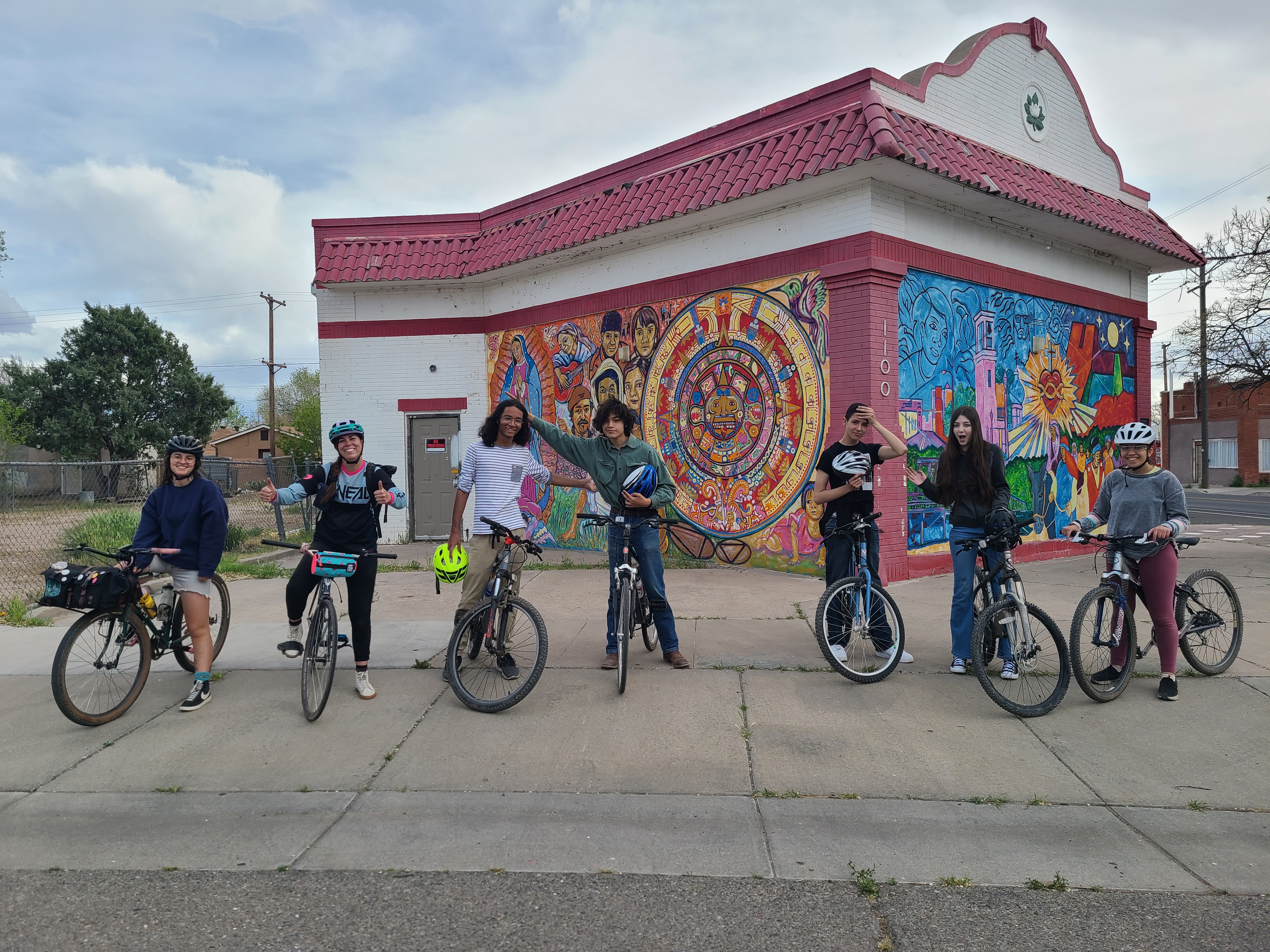 Several young people stand with bikes in front of a brightly painted building, arms in the air